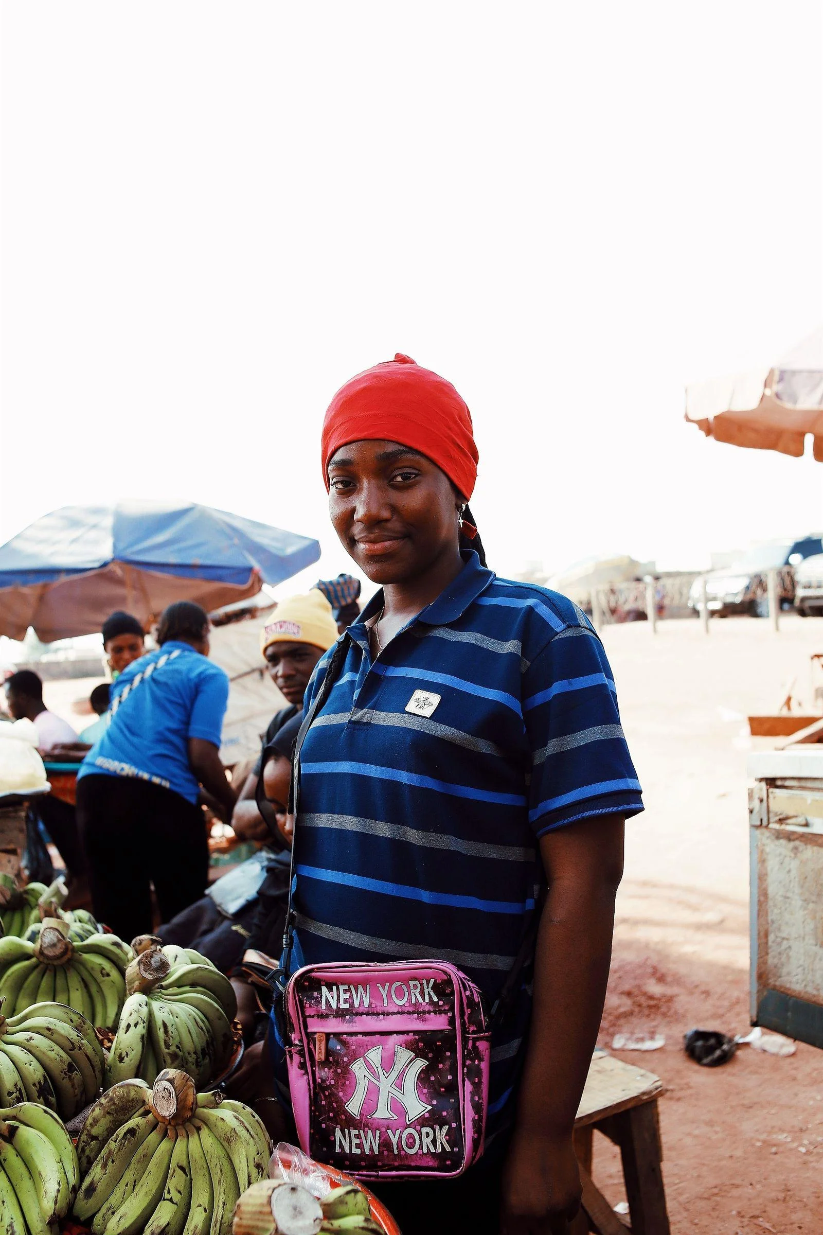 A woman in a red headscarf stands beside plantains at an outdoor Abuja market